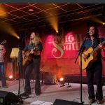 Die drei Musiker von High South - Robert, Jamey und Kevin stehen mit ihren Gitarren auf einer Bühne. Im Hintergrund sieht man eine rote Leinwand mit dem Bandlogo