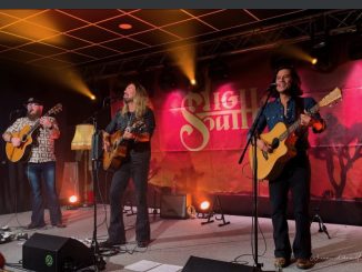 Die drei Musiker von High South - Robert, Jamey und Kevin stehen mit ihren Gitarren auf einer Bühne. Im Hintergrund sieht man eine rote Leinwand mit dem Bandlogo