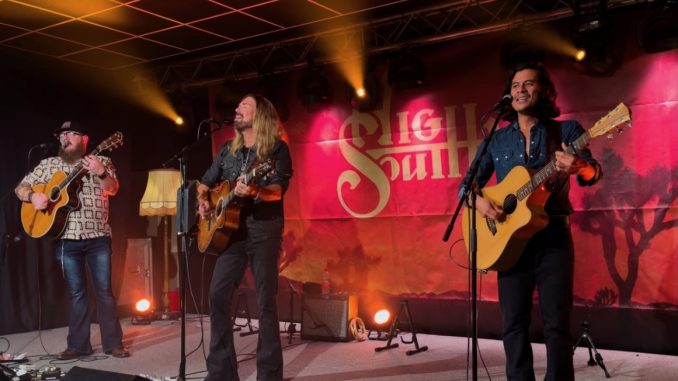 Die drei Musiker von High South - Robert, Jamey und Kevin stehen mit ihren Gitarren auf einer Bühne. Im Hintergrund sieht man eine rote Leinwand mit dem Bandlogo