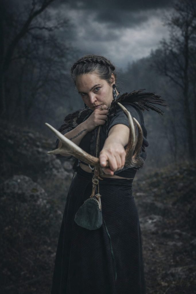 Die Sängerin steht vor einem dunklen Hintergrund, hat ein Geweih in der Hand und zeigt mit diesem in Richtung des Betrachters