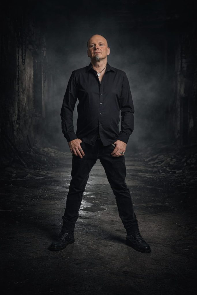 Das Bild zeigt den Bassisten Andi. Er steht, schwarz gekleidet, vor einem dunklen Hintergrund, der einen Weg in einen Wald darstellt.