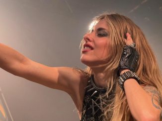 Auf dem Bild sieht man die Sängerin der Metalband Burning Witches wie sie die Hand mit dem Mikro hebt und die andere Hand an ihr Ohr hält