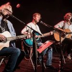 Auf dem Foto sieht man die drei Musiker Ulli Bär, Andy Baum und Matthias Kempf mit ihren Gitarren auf einer Bühne sitzen