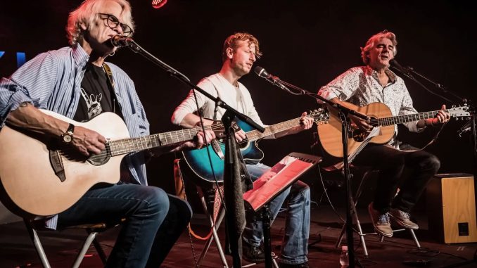 Auf dem Foto sieht man die drei Musiker Ulli Bär, Andy Baum und Matthias Kempf mit ihren Gitarren auf einer Bühne sitzen
