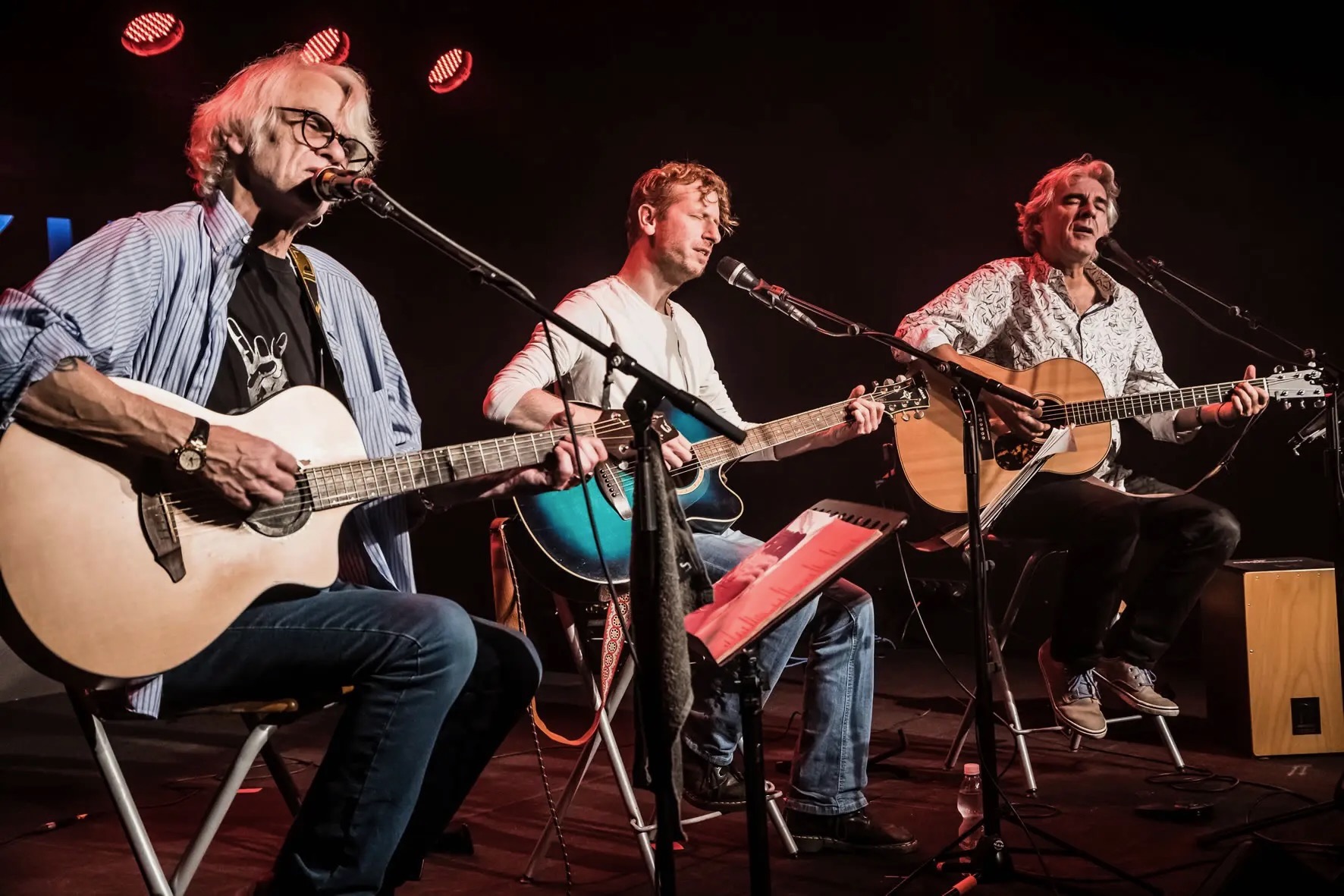 Auf dem Foto sieht man die drei Musiker Ulli Bär, Andy Baum und Matthias Kempf mit ihren Gitarren auf einer Bühne sitzen